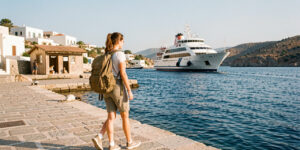 Une jeune bénévole arrive avec un sac à dos sur le quai ensoleillé d’une île grecque, avec un ferry dans le port et un refuge pour chats au loin.