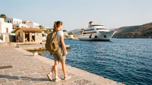 Une jeune bénévole arrive avec un sac à dos sur le quai ensoleillé d’une île grecque, avec un ferry dans le port et un refuge pour chats au loin.